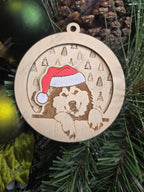 Wooden Christmas ornament with an alaskan malmute wearing a Santa hat, layered on top of a festive pattern, on a green pine branch.