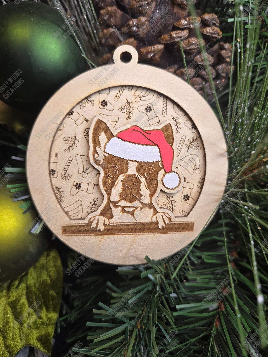 Wooden Christmas ornament with a boston terrier wearing a Santa hat, layered on a festive pattern, on a green Christmas tree.