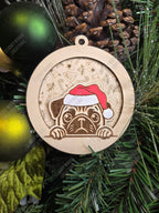 Wooden Christmas ornament with a pug wearing a Santa hat, layered on a festive background piece, on a tree.