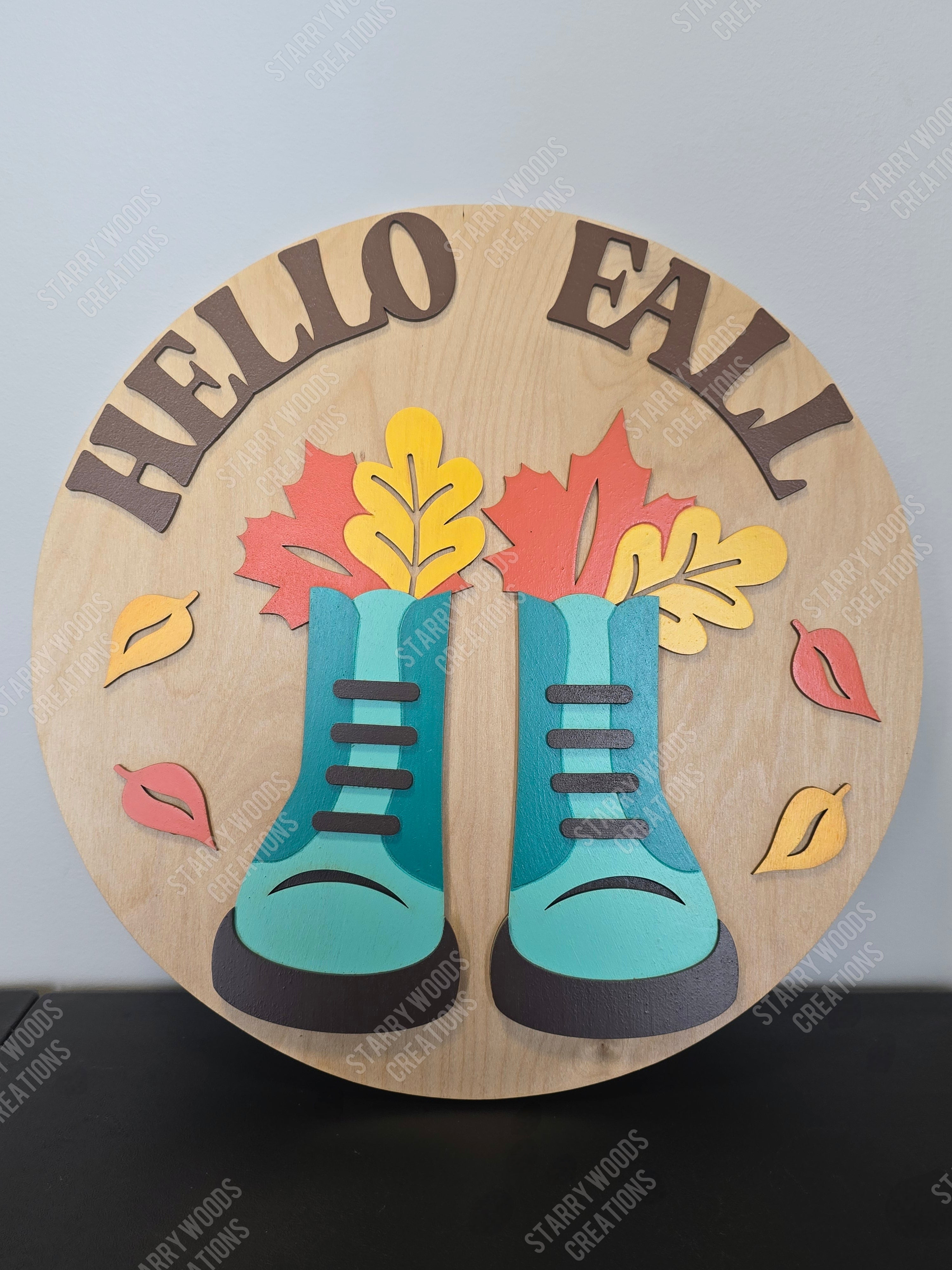 Decorative round sign with blue boots, colorful leaves, and 'Hello Fall' text on a plain background.