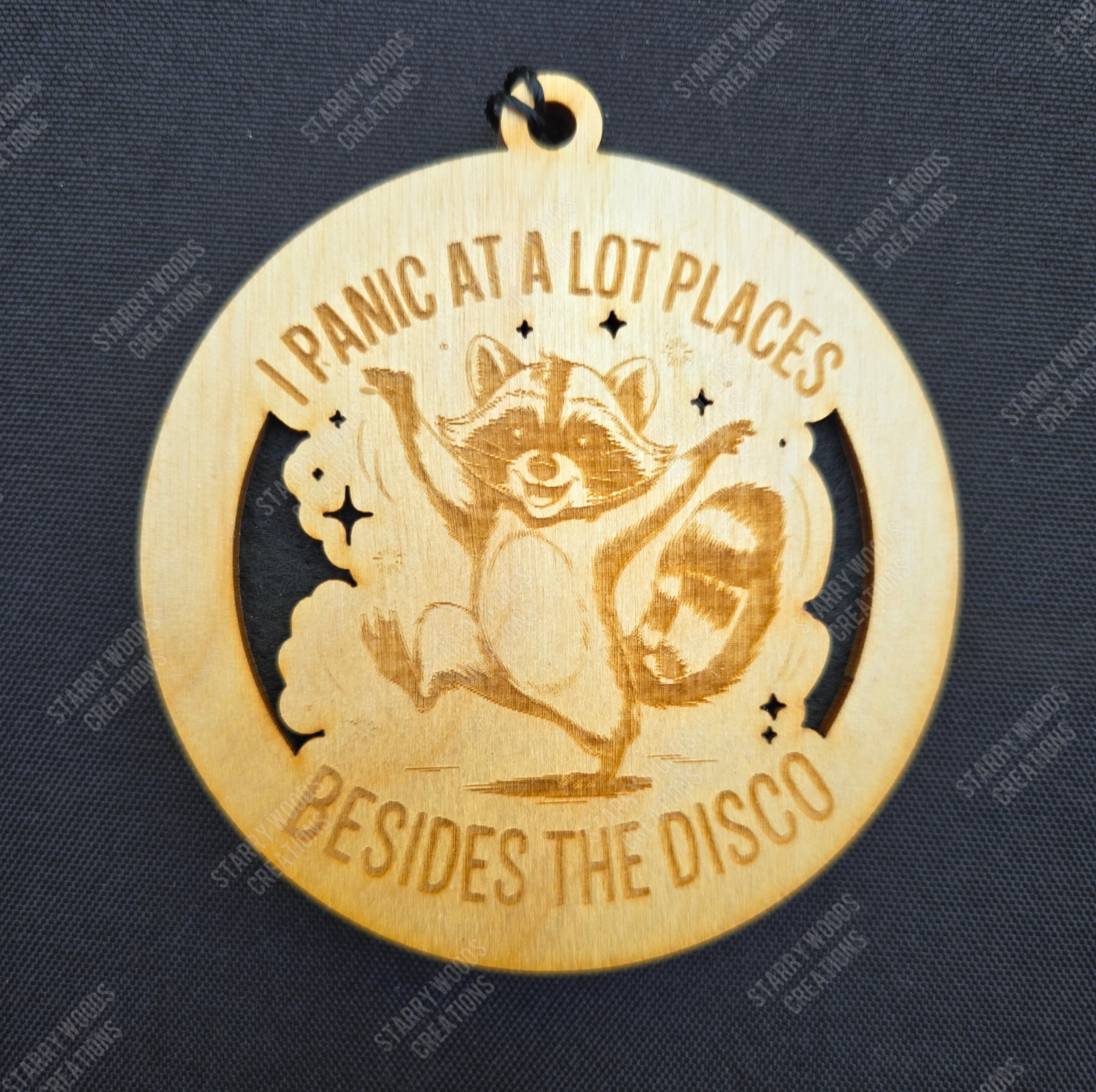 Wooden hanging air freshener with raccoon illustration and "I panic at a lot of places beside the disco" text on a dark blue background